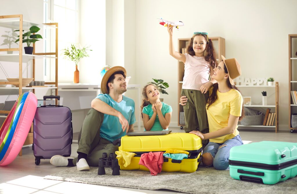 Happy family with children preparing for journey going on adventure trip. Smiling father, mother and sister looking at cheerful daughter playing with toy plane. Home interior with luggage suitcase