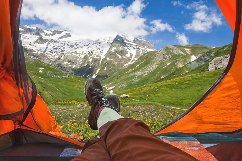 View from orange tent with feet of resting tourist or hicker on snowy mountain peaks and green valleys on sunny day. Alps, France. Lifestyle concept