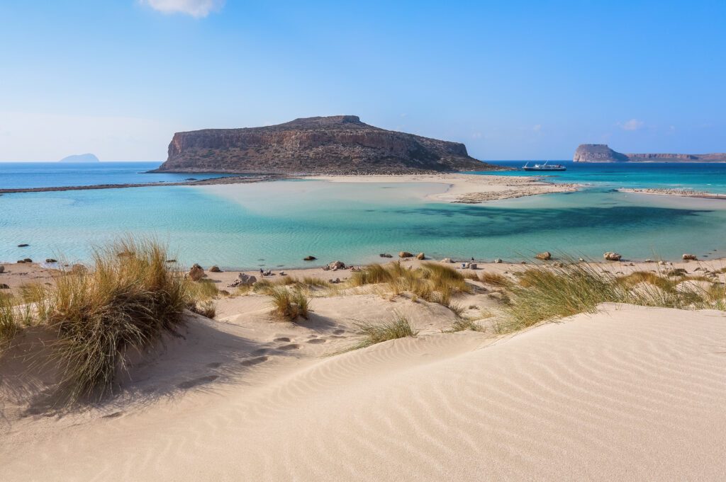 The white sand beside the sea, where the bottom can be seen, the beach with people. Beautiful mountains on a blue horizon. Incredible Balos. A landscape on a summer sunny day. Crete Island, Greece.