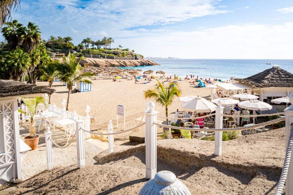 A slice of paradise at the luxurious Playa del Duque beach, with palm trees in the tropical afternoon sunlight and a panoramic view of the sparkling Atlantic Ocean from the Calle el Mirador promenade.