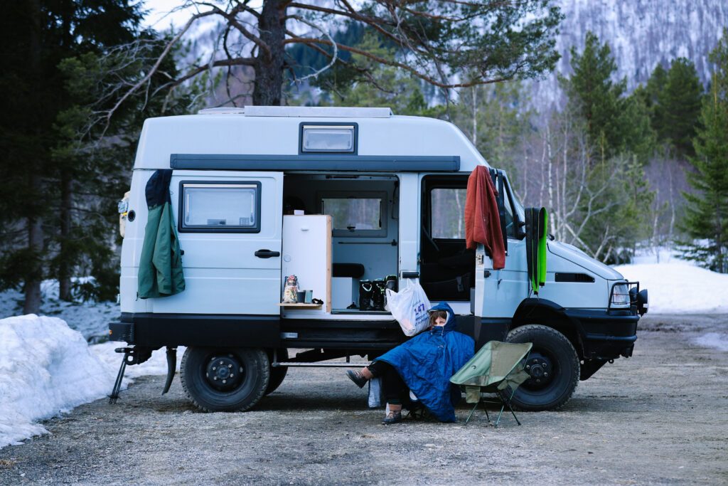 Cosy looking woman in adventure hiking blanket sit on camping chair in front of self converted overlanding 4x4 camping vehicle. Camper vanlife lifestyle. Ski touring adventure in a van