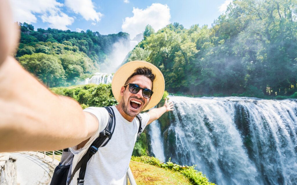 Handsome tourist visiting national park taking selfie picture in front of waterfall - Traveling life style concept with happy man wearing hat and sunglasses enjoying freedom in the nature