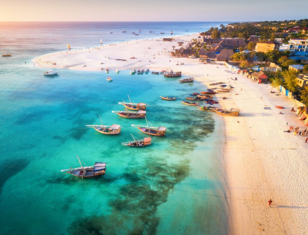 Aerial view of the fishing boats on tropical sea coast with sandy beach at sunset. Summer holiday on Indian Ocean, Zanzibar, Africa. Landscape with boat, buildings, transparent blue water. Top view