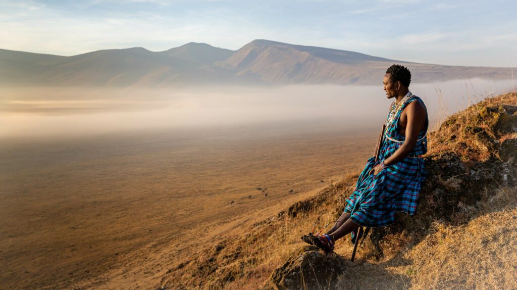 Masai Wwarrior sitting at the edge of one of Ngorongoro craters looking at the horizon and enjoying the surnrize