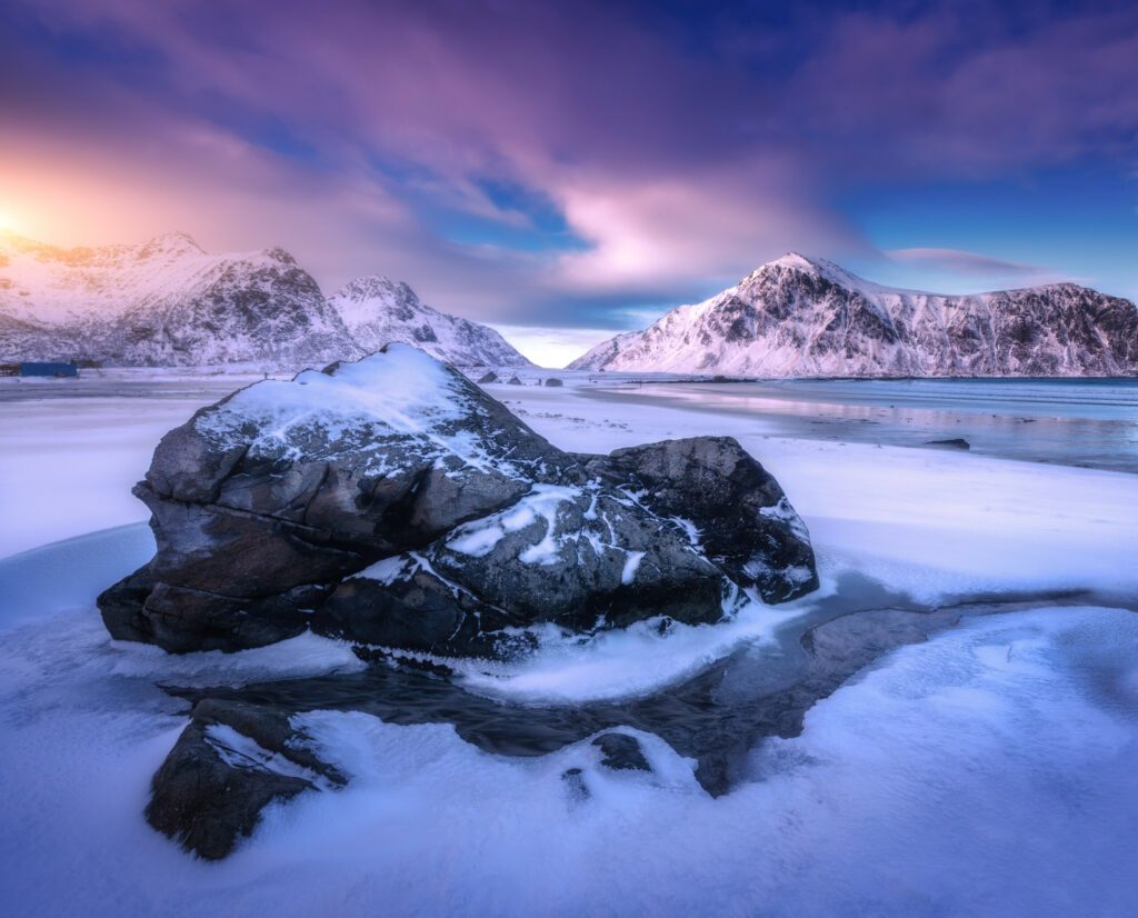 Sandy arctic beach with blue sea, stone in frozen coast and rocks in snow in winter at sunset in Lofoten islands, Norway. Colorful Landscape with snowy mountains, sky with clouds. Norwegian coast
