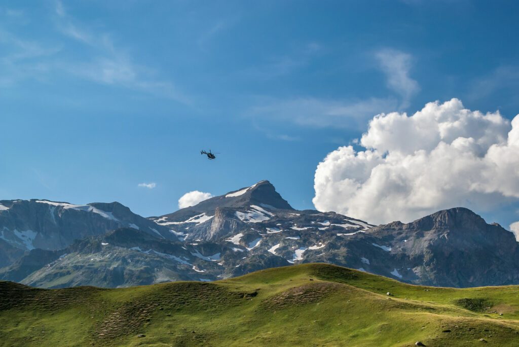 Over a green alpine pasture in a blue sky with white clouds a small black helicopter is flying, French Alps, National Park Vanoise.