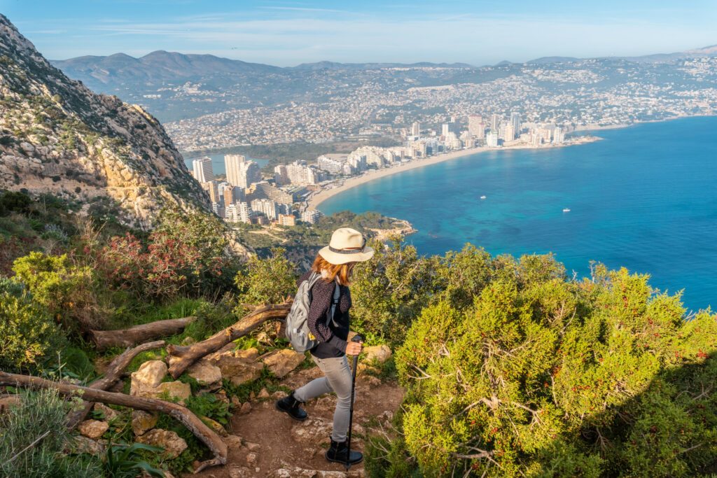 A young hiker wearing a hat on the descent path of the Penon de Ifach Natural Park with the city of Calpe in the background, Valencia. Spain. Mediterranean sea. View of La Fossa beach