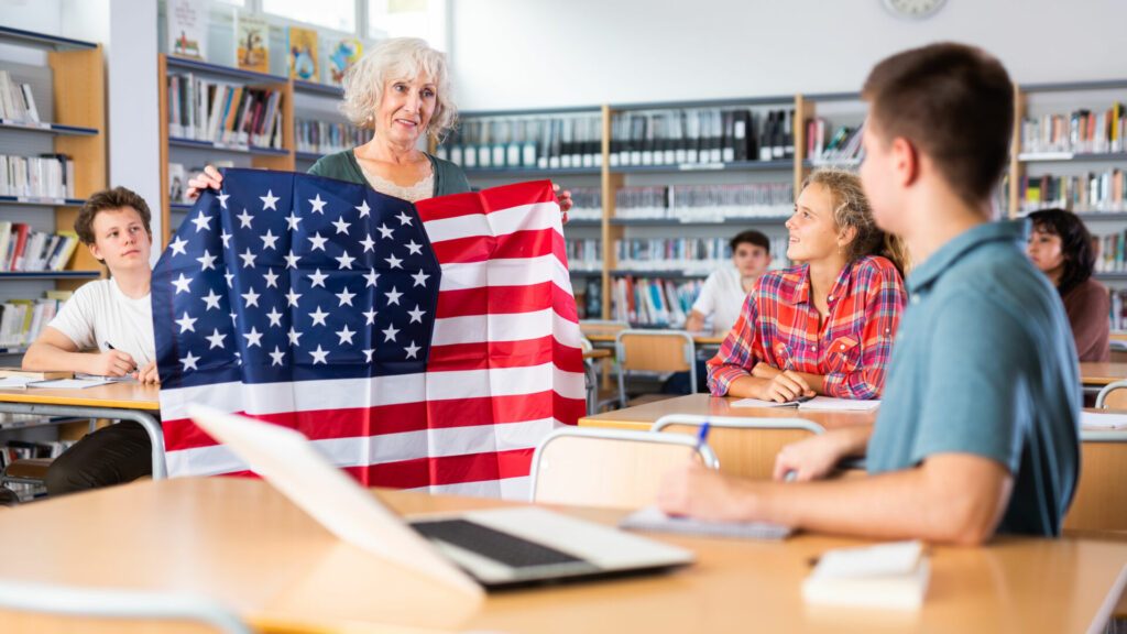 Group of teenagers listening to aged female teacher holding USA flag at Geography lesson in school library