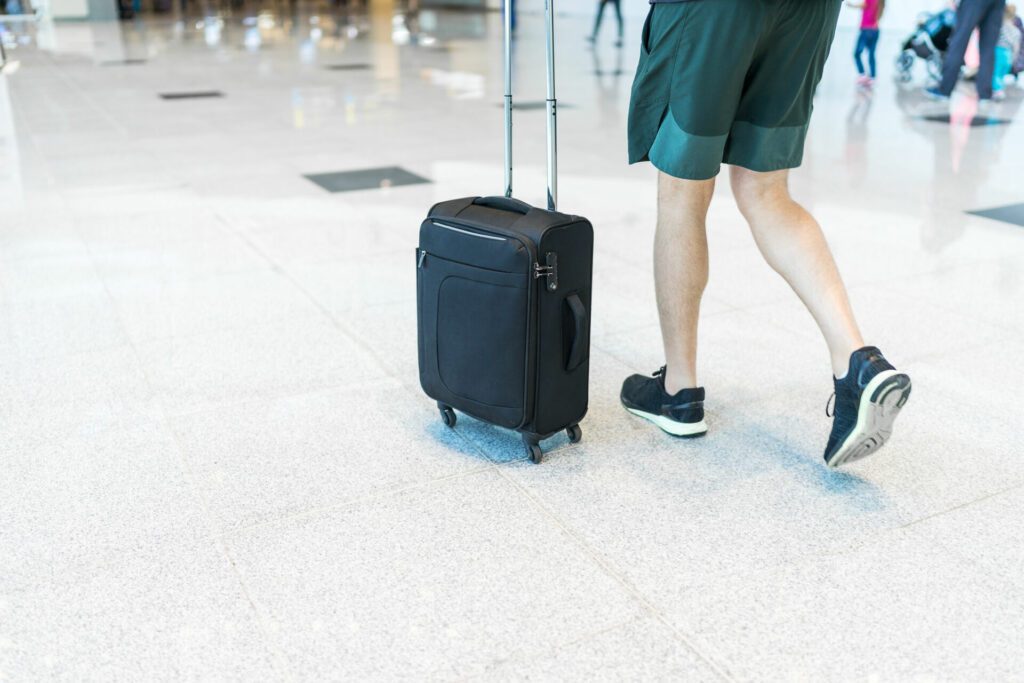 Young man wearing shorts and sneakers is walking through the airport holding a small-sized black suitcase with 4 wheels